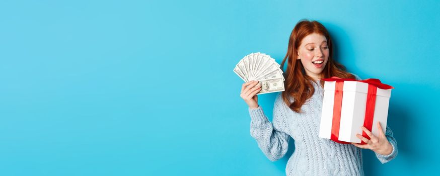 Christmas and shopping concept. Cheerful girl with red hair, holding money and big New Year gift, smiling happy, standing over blue background.