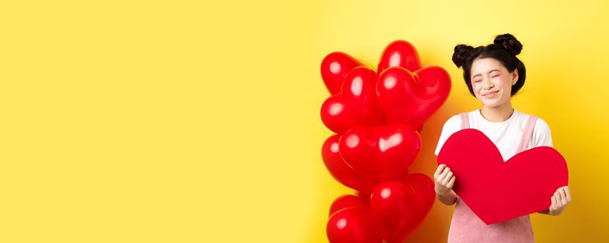 Happy Valentines day. Silly and beautiful asian woman smiling dreamy, showing red heart, imaging romantic date with lover, standing on yellow background.