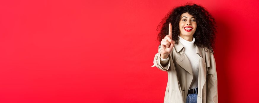 Cheerful woman in trench coat, showing number one finger and smiling, standing on red background.