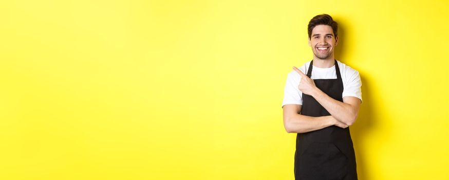 Happy barista pointing finger left and smiling, wearing black apron uniform, standing against yellow background. Copy space