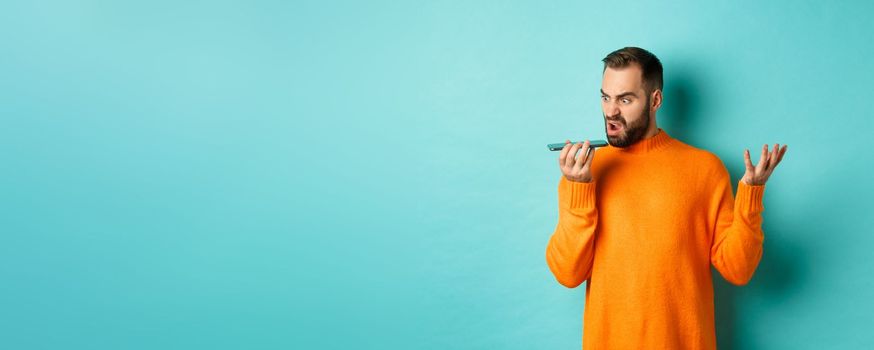 Angry man arguing on speakerphone, record voice message with mad face, standing over light blue background in orange sweater.