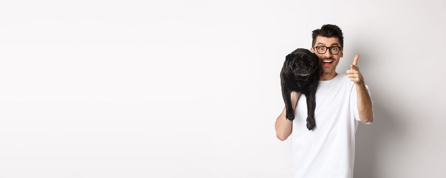 Happy young man holding cute black dog on shoulder and pointing at camera. Hipster guy carry pug on shoulder and staring at camera excited, standing over white background.
