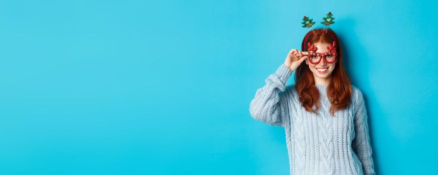 Winter holidays and Christmas sales concept. Beautiful redhead female model celebrating New Year, wearing funny party headband and glasses, smiling at camera.