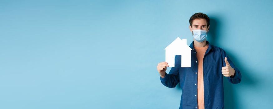 Real estate and covid concept. Young man in face mask showing paper house cutout and thumb-up, recommending agency, standing on blue background.