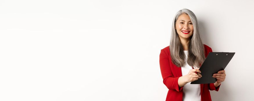 Elegant asian senior businesswoman working with documents, holding pen and clipboard, signing contract and smiling, standing over white background.