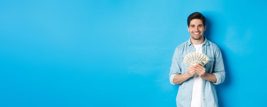 Handsome smiling man holding money, concept of finance and banking, standing over blue background.