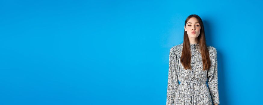 Valentines day and relationship concept. Lovely woman in dress pucker lips for kiss, looking with love at at camera, standing on blue background.