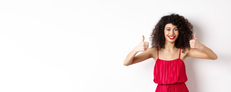 Attractive young woman recommending promo offer, showing thumb up and smiling, like product, standing in festive red dress on white background.