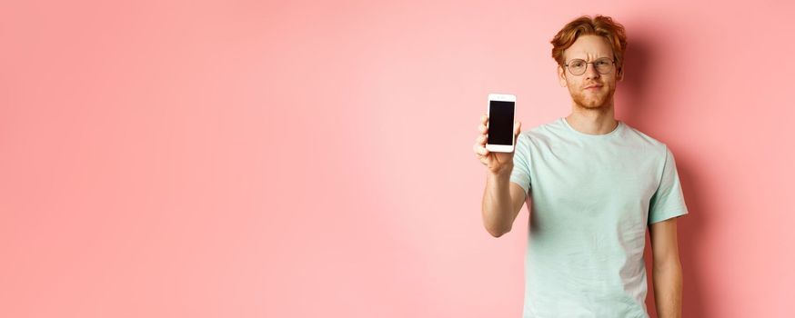 Disappointed male model frowning, showing smartphone screen, standing over pink background.