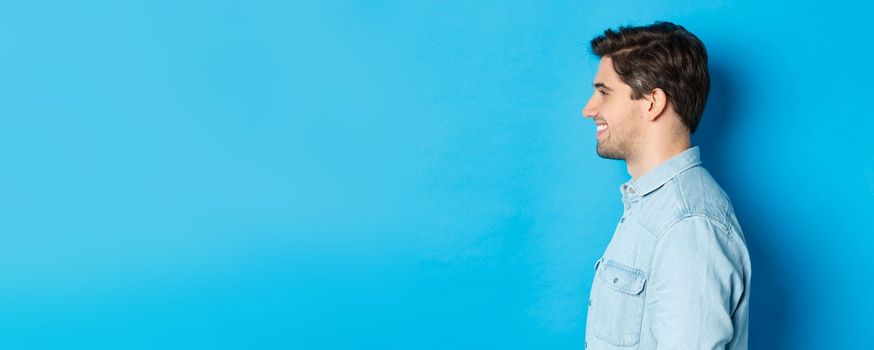 Profile of handsome young man looking left, smiling happy, standing over blue background.