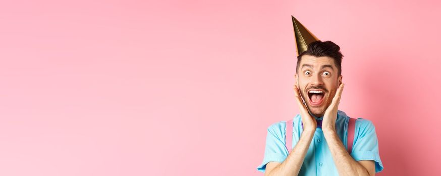 Holidays and celebration concept. Close-up of funny guy in birthday hat shouting surprised, screaming with joy, receiving awesome gift, standing happy on pink background.