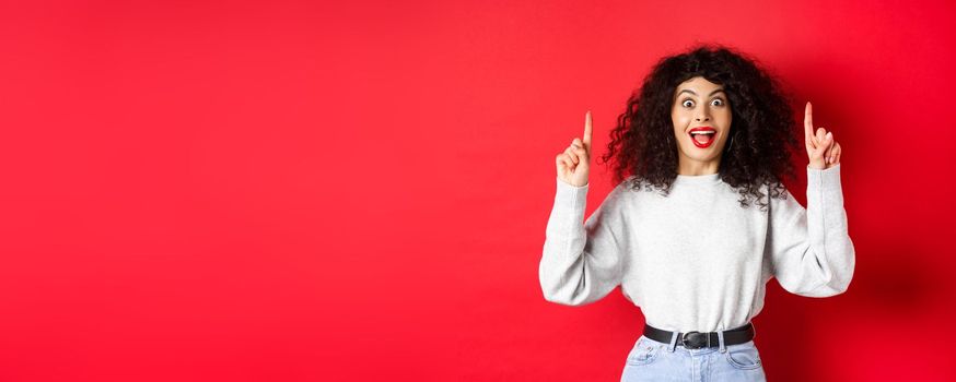 Excited modern girl showing promo, pointing fingers up and smiling amazed, telling big news, standing on red background.