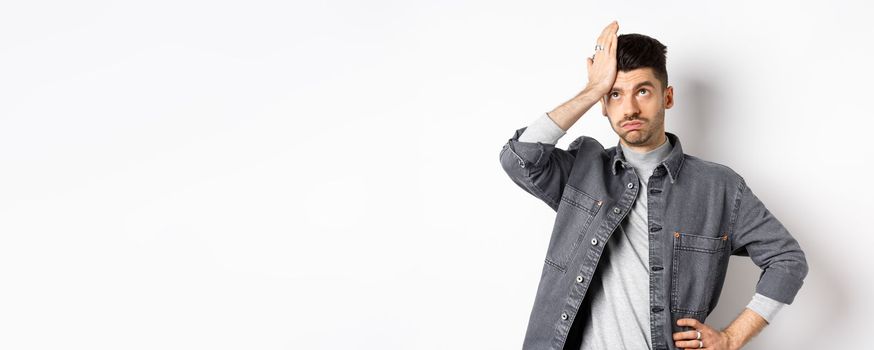 Annoyed young man looking tired or bothered, roll eyes and slap forehead, making facepalm from something stupid, standing on white background.