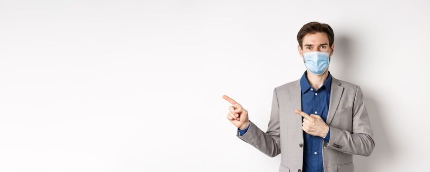 Smiling businessman in suit and medical mask showing advertisement, pointing fingers left and looking friendly, white background. Covid-19 and pandemic concept.