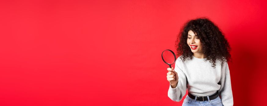 Young woman looking through magnifying glass, investigating or searching for something, stare curious aside, found interesting thing, standing on red background.