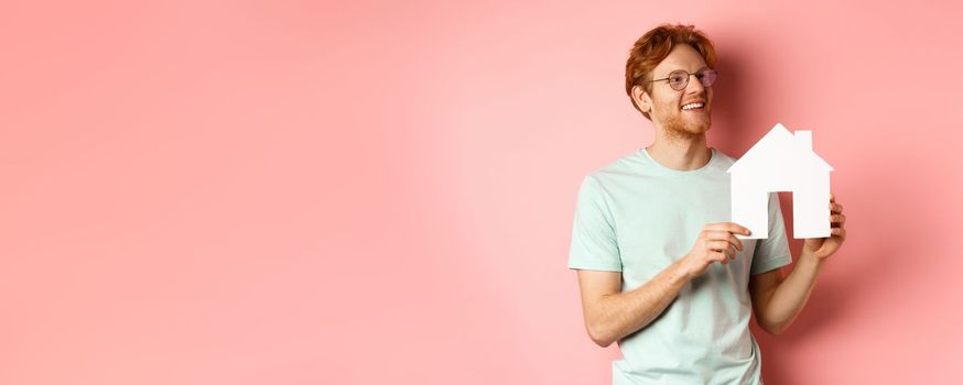 Real estate. Happy redhead man dreaming of buying property, looking right and showing paper house cutout, standing over pink background.