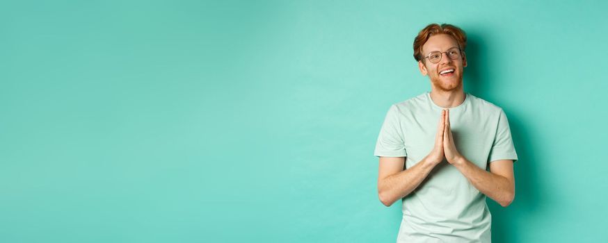 Hopeful redhead man with beard, wearing glasses and t-shirt, holding hands in namaste or plead gesture and looking right, smiling and thanking, standing over turquoise background.