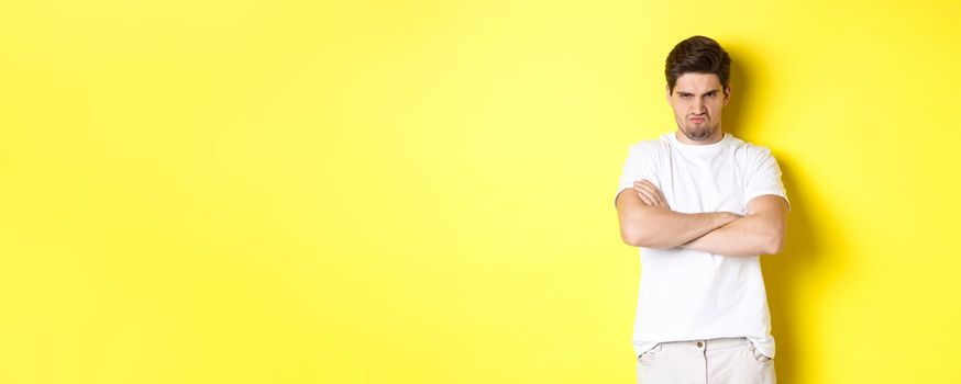 Offended and mad guy, frowning and sulking, holding hands crossed on chest, standing angry against yellow background.