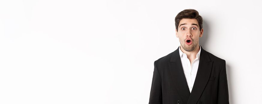 Close-up of attractive man in black suit, looking surprised and impressed at advertisement, standing over white background.