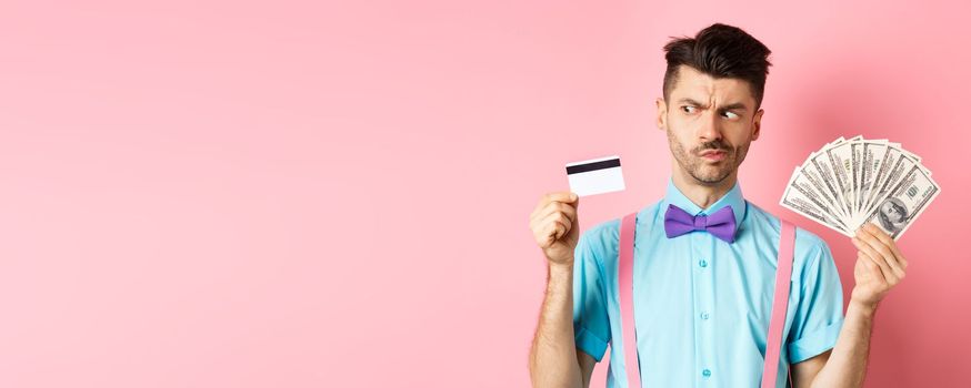 Doubtful and indecisive guy looking hesitant at plastic credit card, holding cash dollars, standing over pink background.