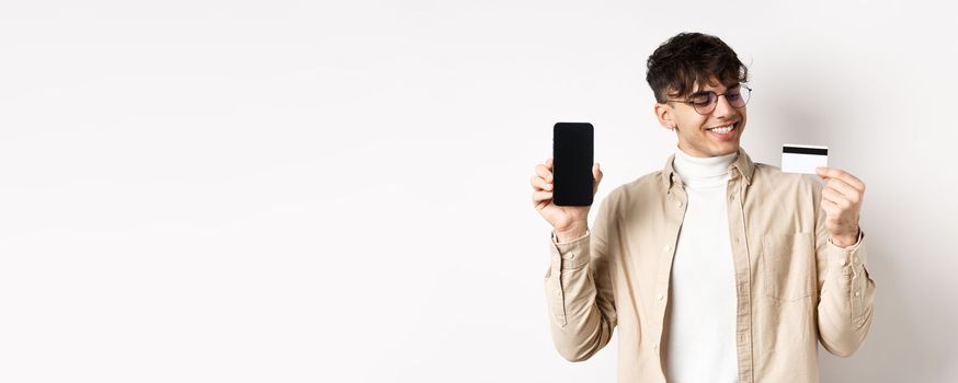 Satisfied young man looking pleased at credit card, showing empty smartphone screen, standing on white background.