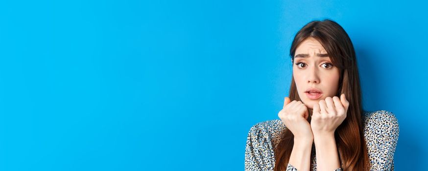 Close-up of scared timid girl looking afraid, shaking from fear, press hands to body and staring frightened, standing on blue background.