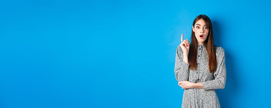 Excited female model in dress raising finger, pitching idea and look amazed at camera, found solution or have plan, understood something, standing on blue background.