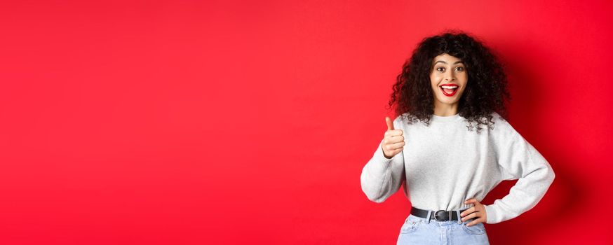 Happy young woman with curly hair praising good work, say well done and show thumb up gesture, approve and praise you, standing on red background.