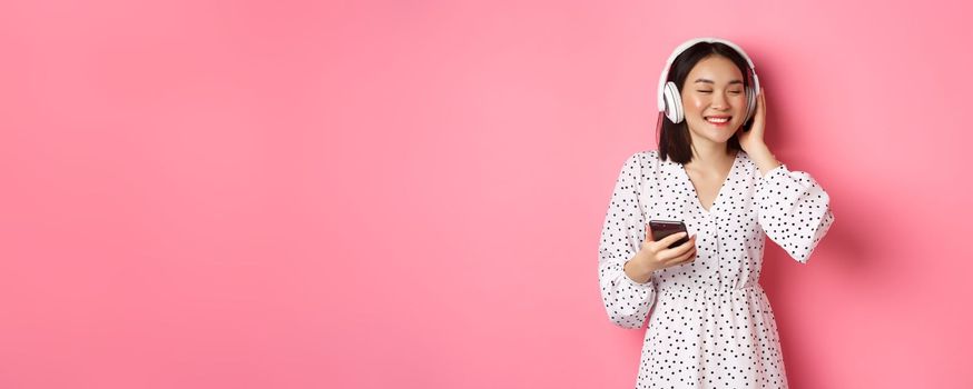 Romantic asian girl listening music in headphones, smiling with closed eyes, holding mobile phone, standing over pink background.