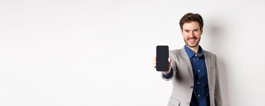 E-commerce and online shopping concept. Confident businessman in suit stretch out hand with empty smartphone screen, showing on phone, standing against white background.