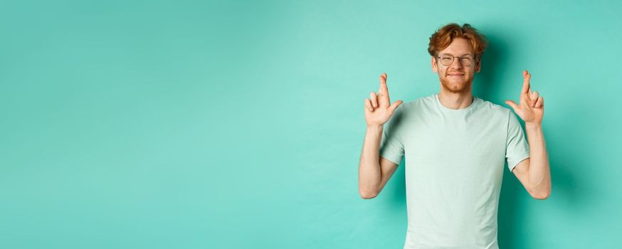 Young cheerful guy with red hair and beard, wearing glasses, smiling and cross fingers for good luck, making wish and looking optimistic, standing over mint background.