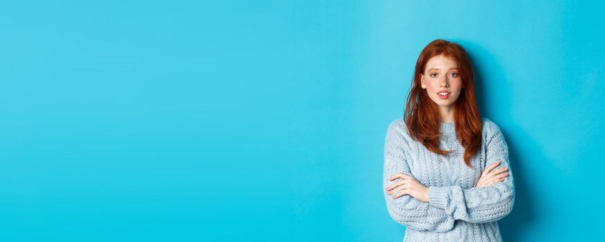 Redhead teenage girl in warm sweater standing over blue background, cross arms on chest with confidence.