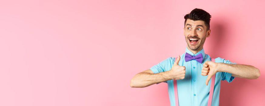 Cheerful man looking happy left, showing thumbs up down, judging product, give positive and negative feedback, standing over pink background.