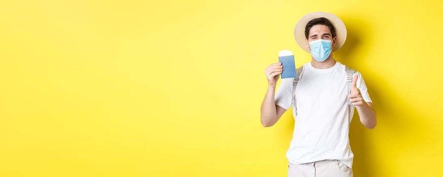 Concept of covid-19, tourism and pandemic. Happy guy tourist in medical mask showing passport with tickets for vacation, winking and pointing at camera, yellow background.