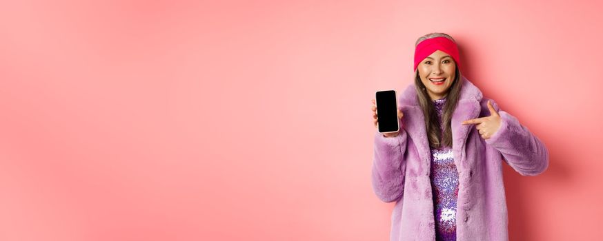 Online shopping and fashion concept. Stylish asian mature lady showing blank smartphone screen, pointing at phone and smiling, pink background.