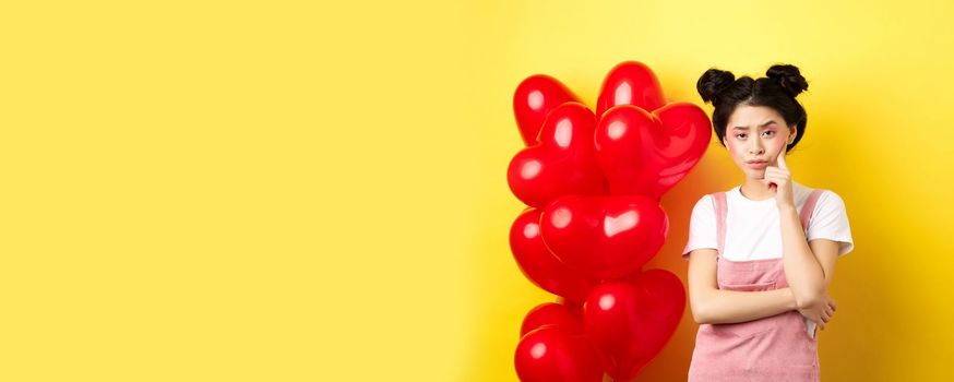 Valentines day concept. Unamused and bored asian girlfriend staring skeptical at camera, waiting for something near red hearts balloons, yellow background.