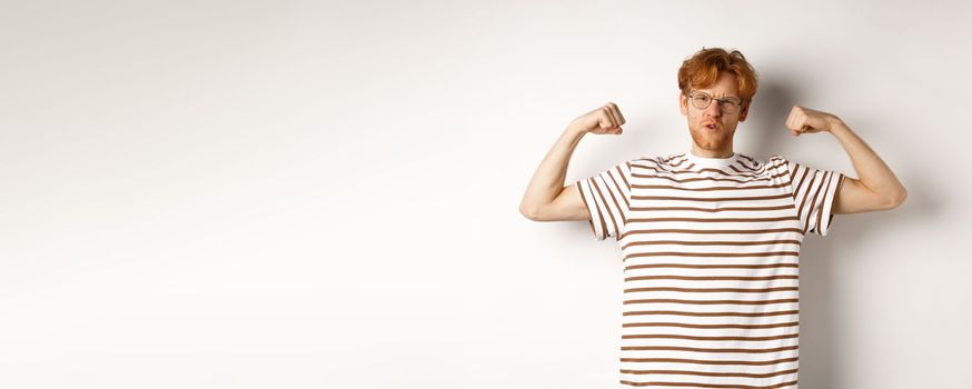 Image of confident and strong redhead man flexing biceps, showing muscles after gym, standing over white background.