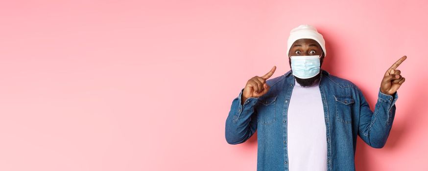Covid-19, lifestyle and lockdown concept. Impressed Black man in face mask showing announcement, pointing right at promo and staring at camera amazed, pink background.