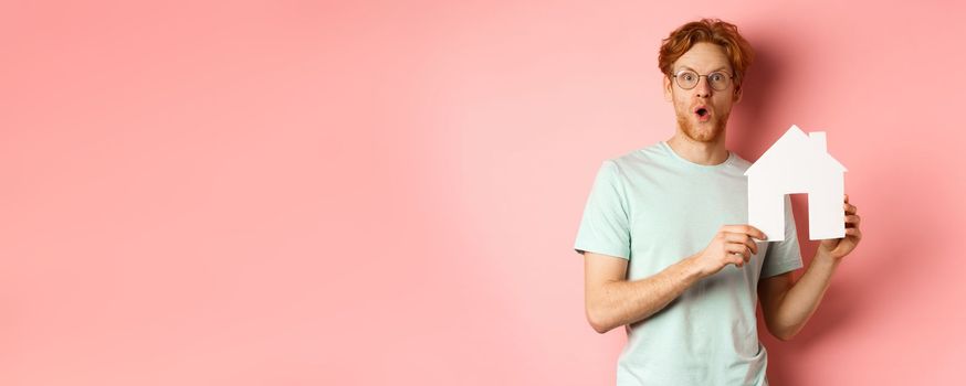 Real estate. Surprised young man with red hair and beard, wearing glasses and t-shirt, showing paper house cutout and looking impressed, saying wow, standing over pink background.
