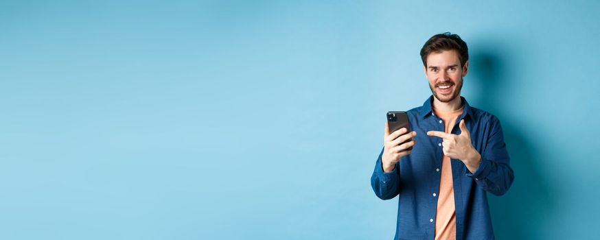 Handsome smiling man pointing finger at smartphone and looking pleased at camera, recommending app, standing on blue background.