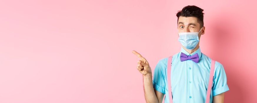 Covid-19, pandemic and health concept. Cheerful caucasian man in face mask winking at camera, showing good deal, pointing finger left at logo, pink background.