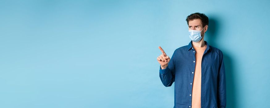 Covid-19 and healthcare concept. Confused and doubtful man in face mask frowning, pointing and looking at empty space, standing on blue background.