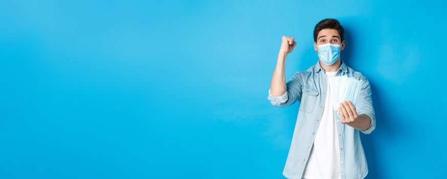 Concept of coronavirus, quarantine and social distancing. Cheerful man showing medical masks and making fist pump, celebrating or triumphing, standing against blue background.