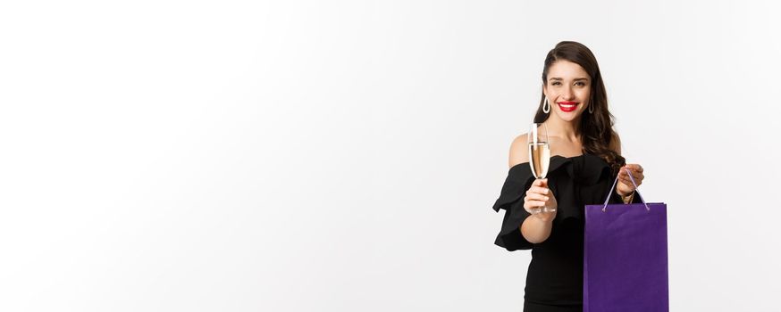 Beautiful and stylish brunette woman raising glass of champagne, celebrating christmas, holding shopping bag with presents, standing over white background.