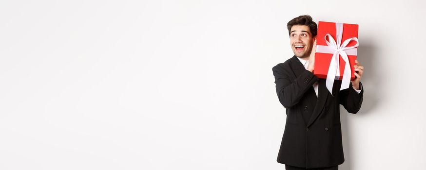 Image of handsome dreamy guy in black suit, shaking box with present to wonder whats inside, standing against white background happy.