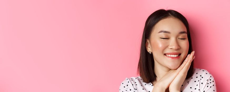 Beauty and skin care concept. Headshot of adorable and dreamy asian woman close eyes, smiling nostalgic, standing against pink background.