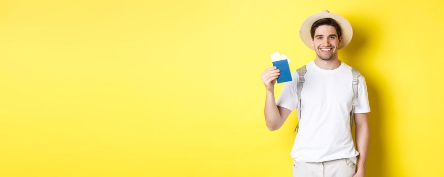 Tourism and vacation. Young smiling tourist showing passport with tickets, going on a trip, standing against yellow background.