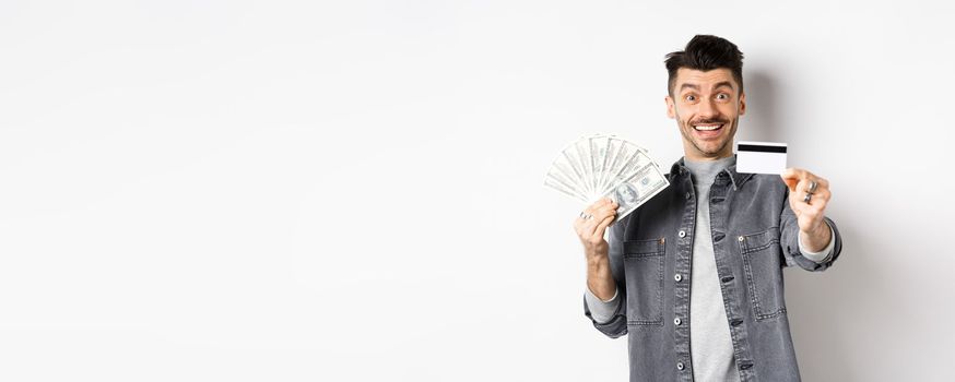 Image of handsome guy holding dollar bills but suggesting use plastic credit card, smiling friendly at camera, standing on white background.