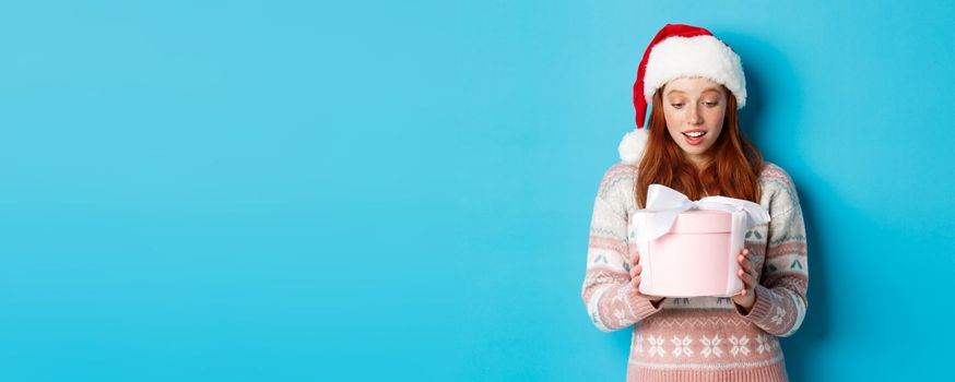 Winter and Christmas Eve concept. Touched and flattered redhead girl looking at box with xmas gift, smiling amazed, standing in santa hat against blue background.