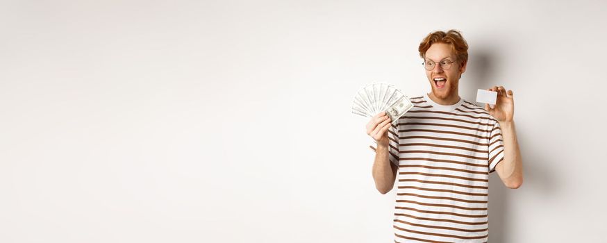 Shopping and finance concept. Cheerful handsome redhead man showing plastic credit card and cash, smiling amazed, white background.